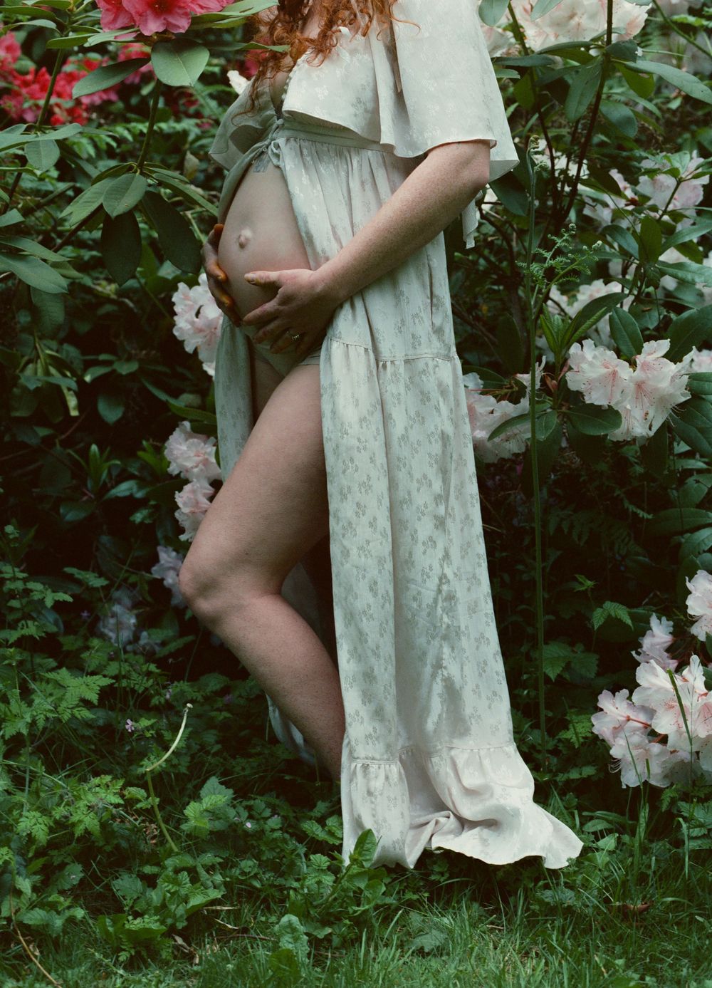 Close-up of pregnant woman in floral maxi dress holding baby bump surrounded by blooming flowers.