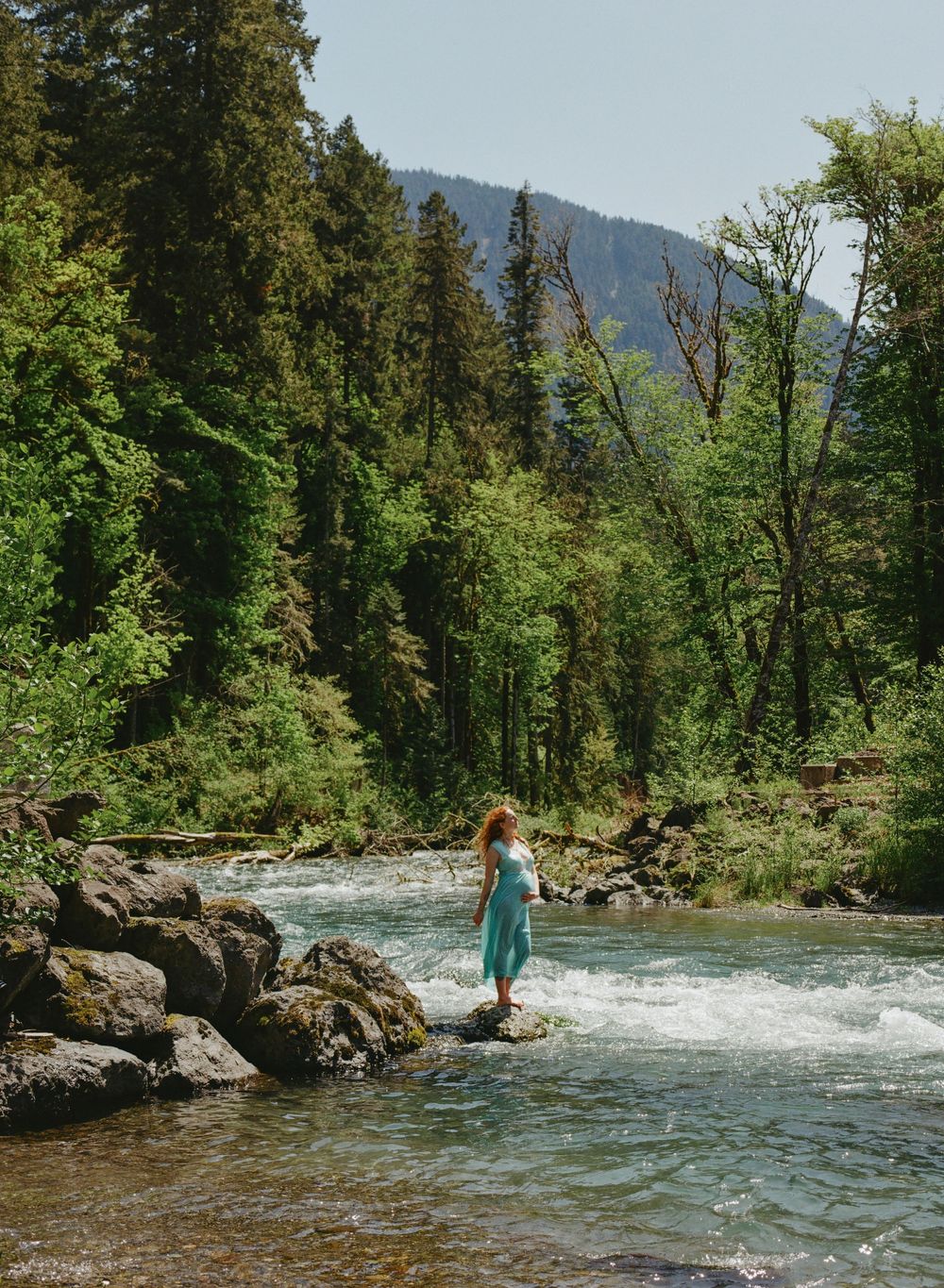 Pregnant woman in teal dress standing on rocks in a mountain river surrounded by forest.