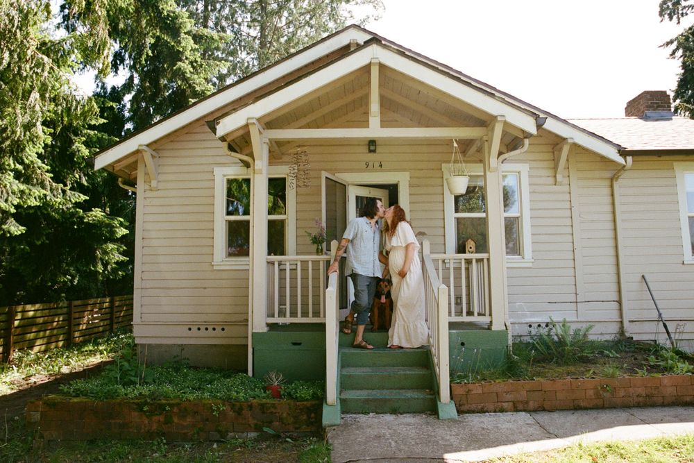 Couple standing on the porch of a charming beige craftsman cottage home surrounded by lush green trees.