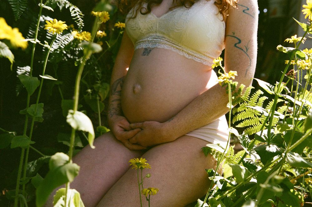 Close-up of a pregnant woman in yellow lingerie cradling her bare baby bump among wildflowers.