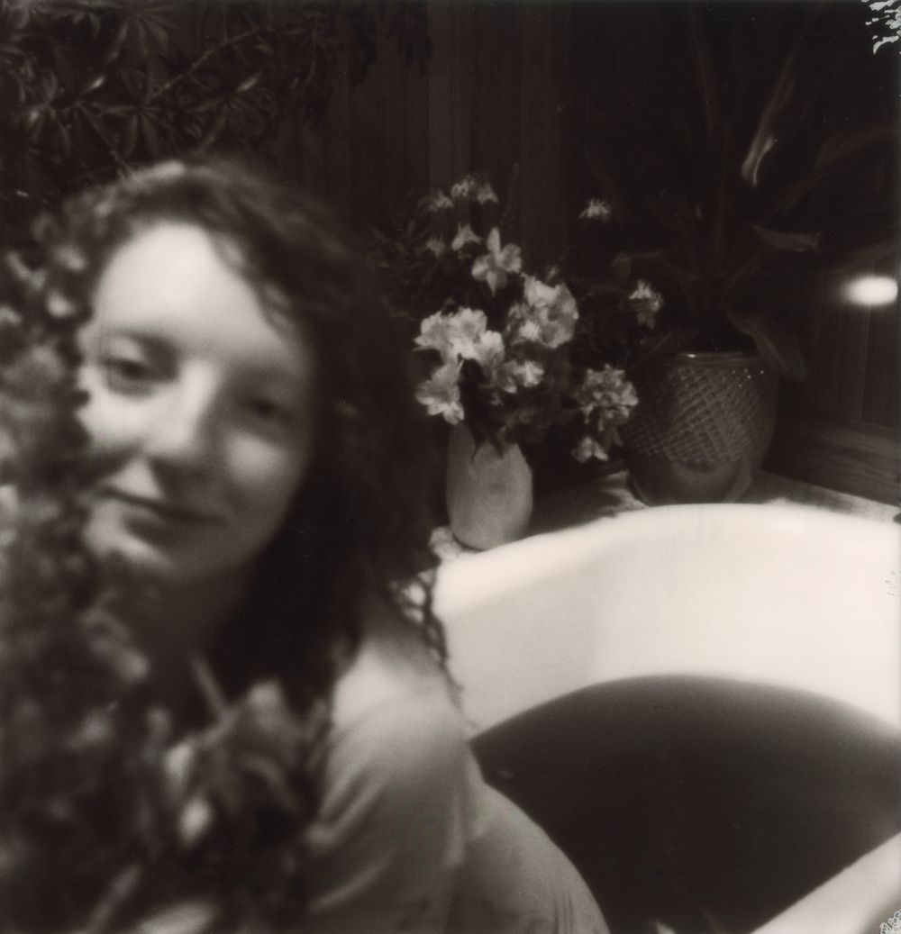 Black and white photo of a young woman with curly hair near flowers and a sink with a vase of blooms.