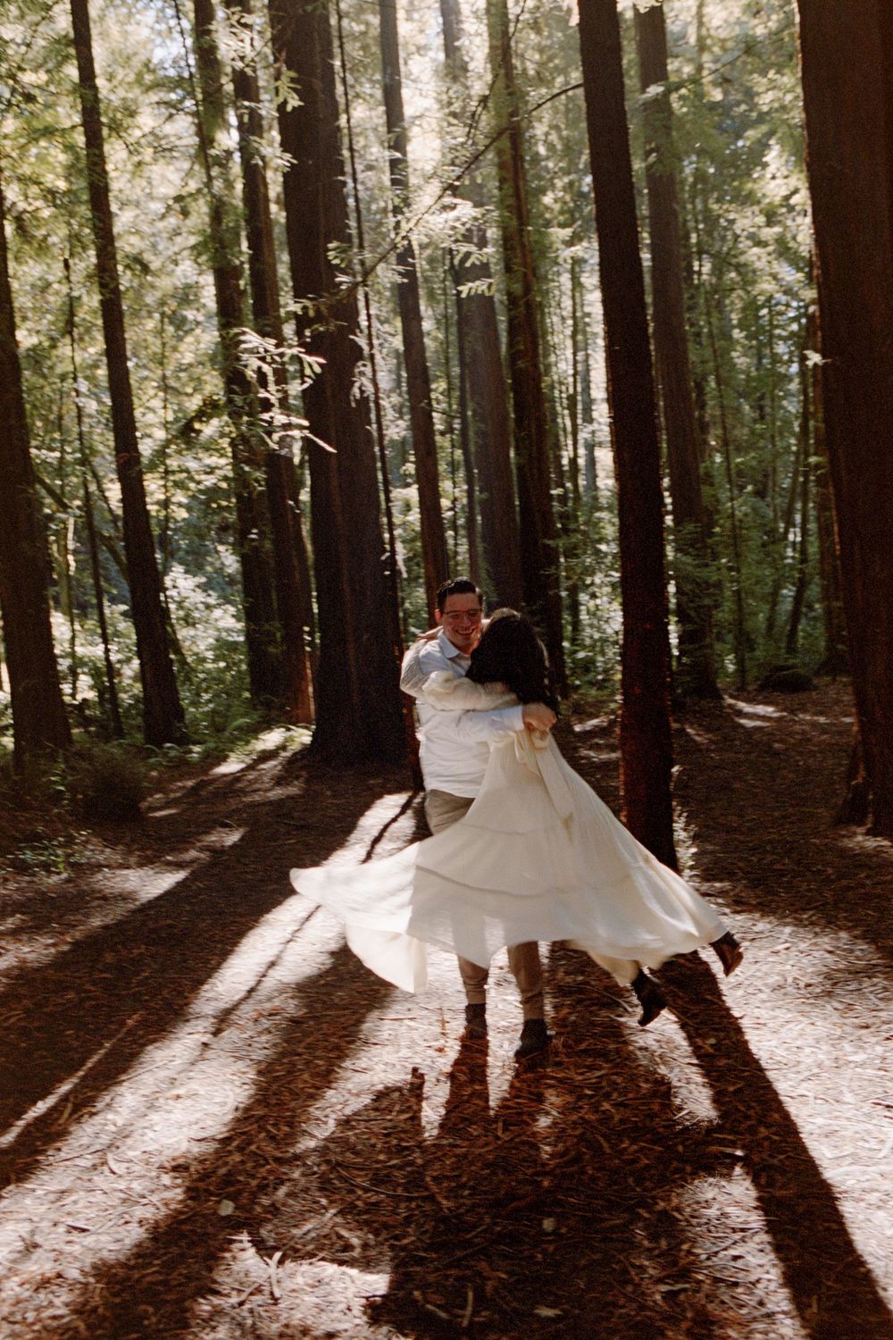 A couple dances joyfully in a sunlit redwood forest, the bride's white dress swirling as they embrace among tall trees.