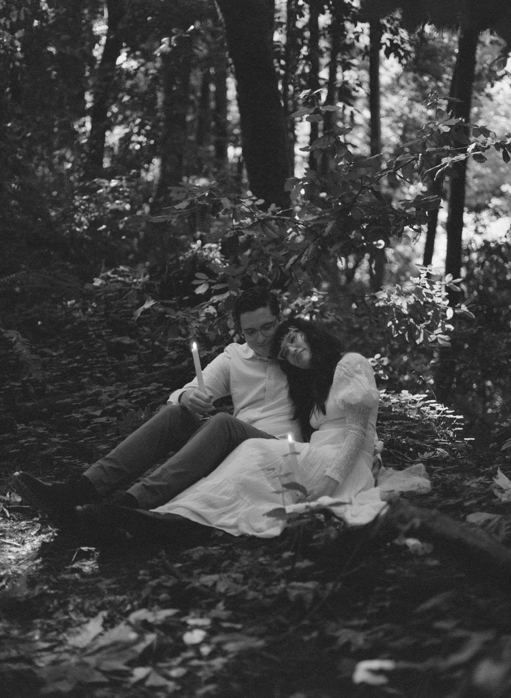 A couple sits together in a forest, leaning on each other in a romantic black and white photo.