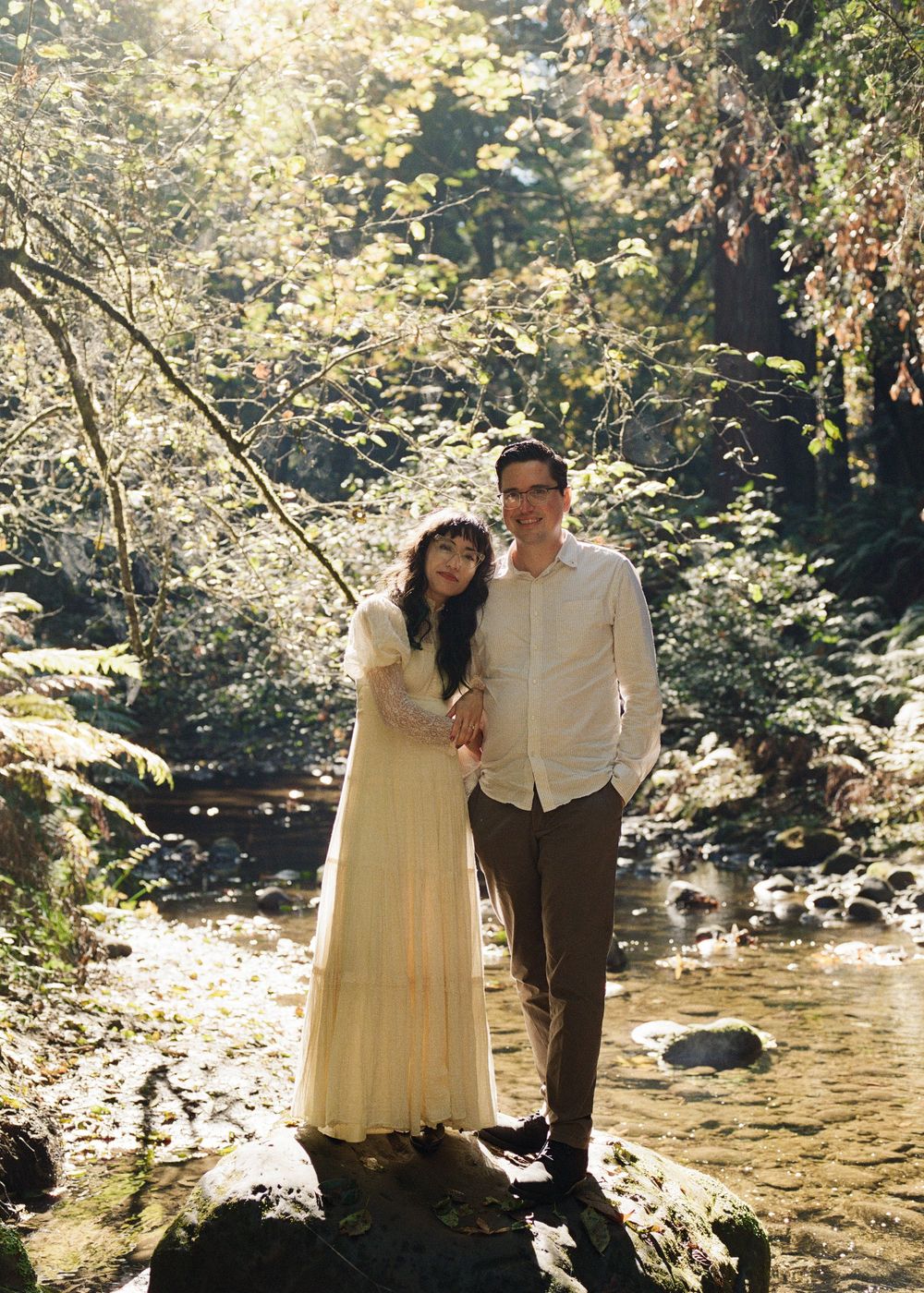 Couple posing together on rocks by a creek surrounded by lush green forest trees in soft natural sunlight.