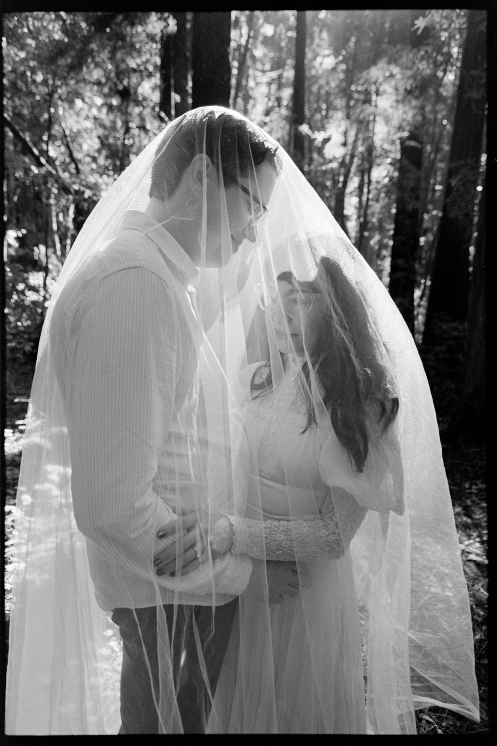 Black and white photo of bride and groom sharing a kiss under a flowing wedding veil in a forest.