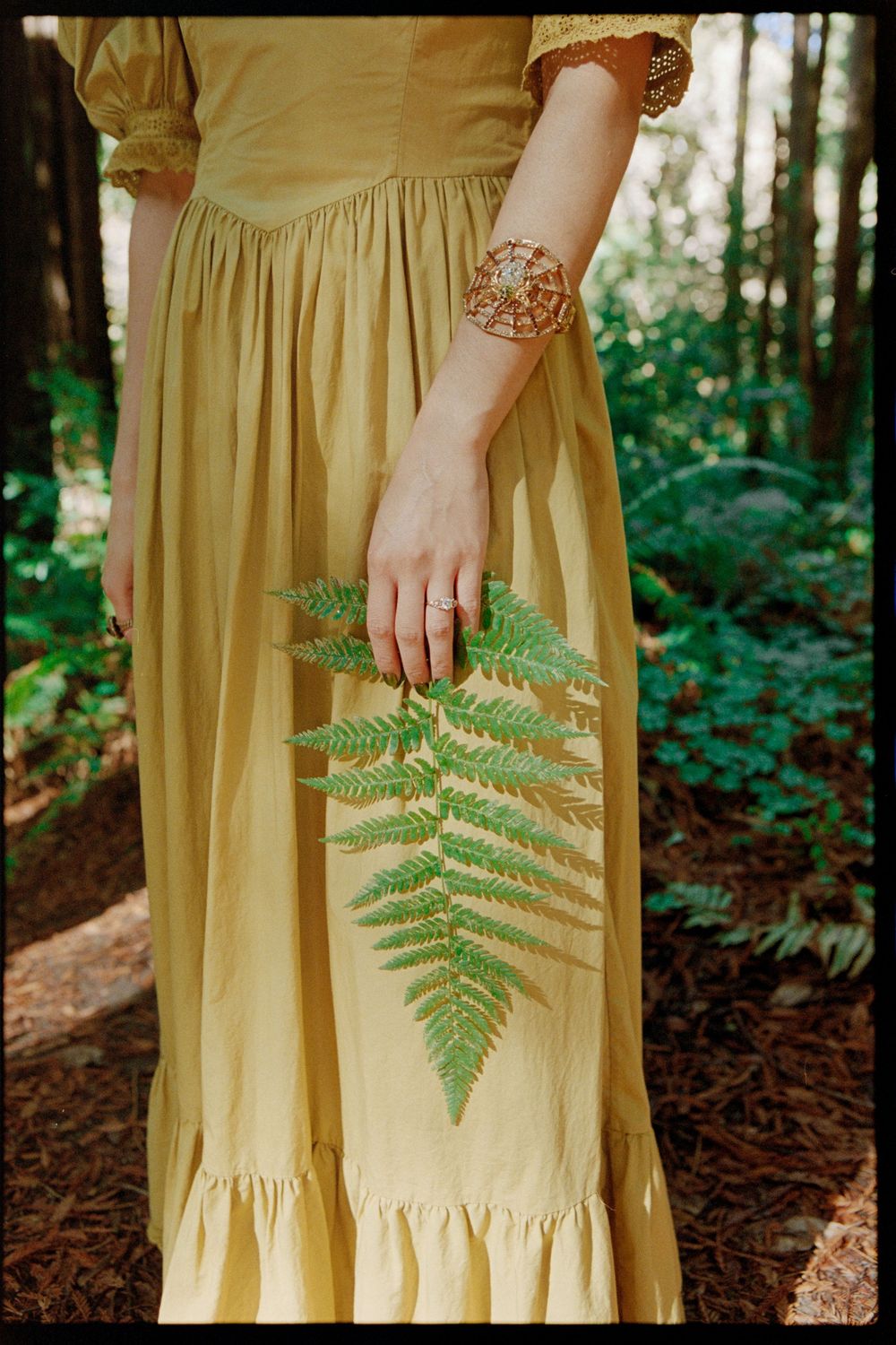 Woman in yellow maxi dress holding a green fern leaf in a forest setting, wearing a decorative bracelet.