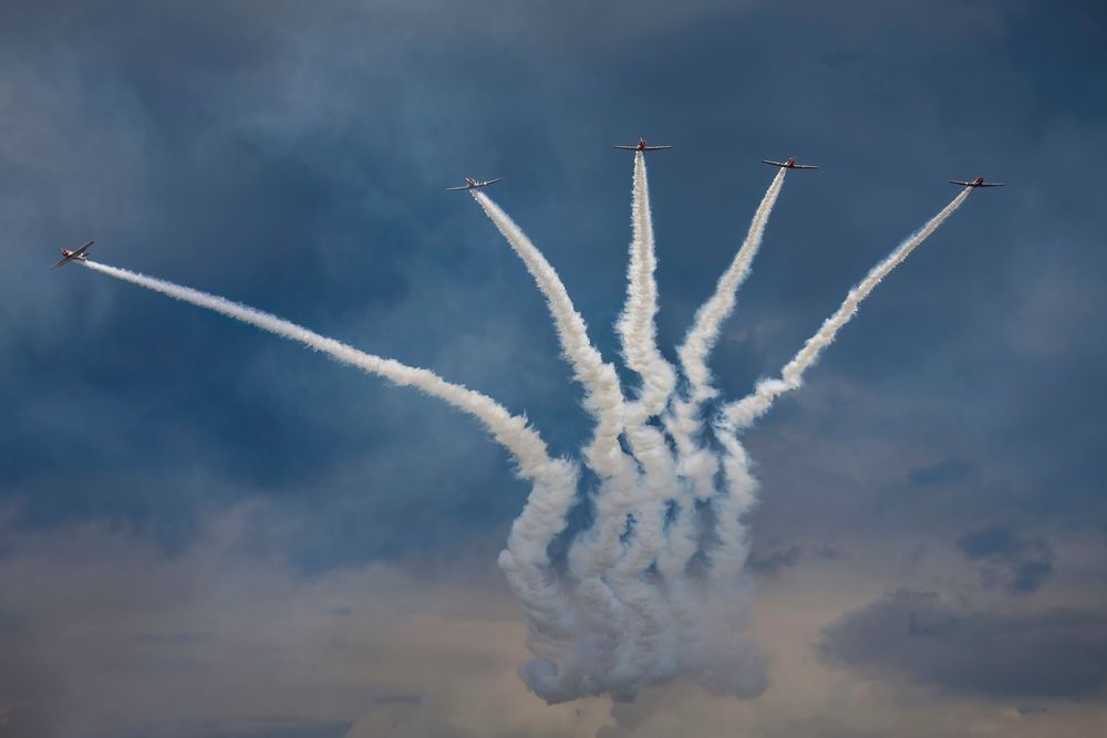 Thunderbirds military jets perform a synchronized aerial maneuver leaving white smoke trails against a cloudy blue sky.
