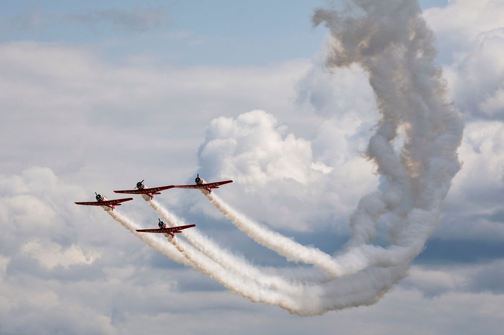 Stunt planes perform synchronized aerial maneuvers leaving white smoke trails against a cloudy blue sky.