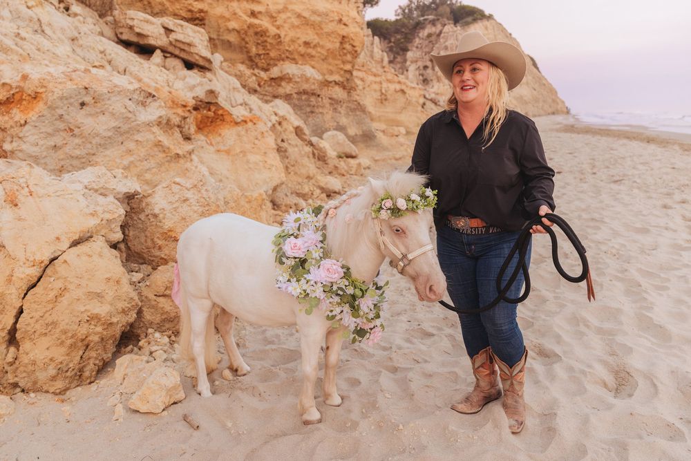 Woman in cowboy hat and boots stands with a flower-adorned white miniature horse on a sandy beach.