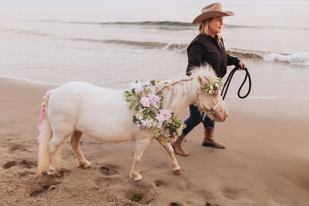 A woman in a cowboy hat leads a decorated white pony with flowers on a sandy beach near the ocean.