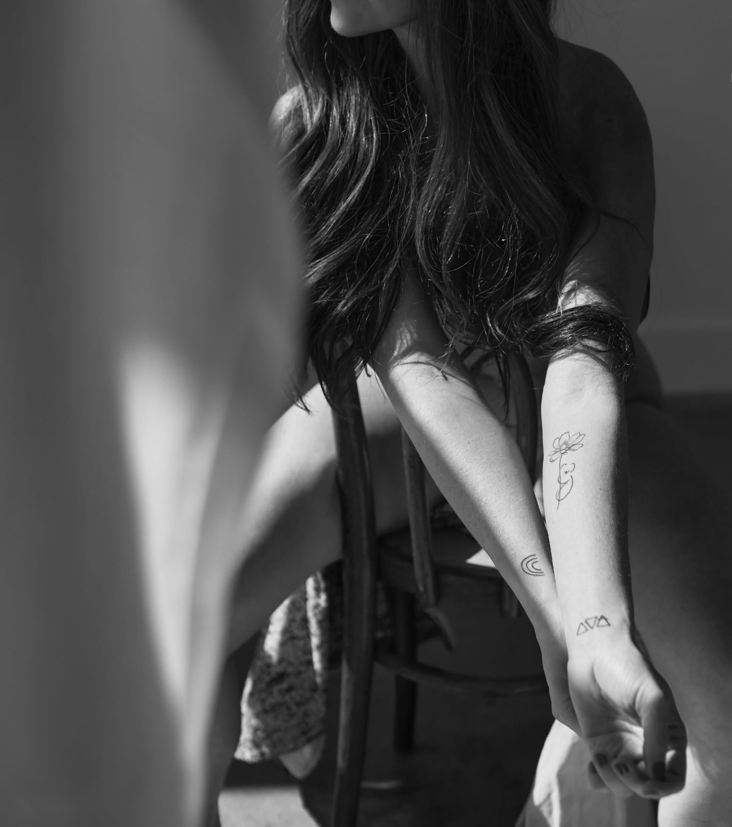 Black and white photo of a woman with long dark hair showing small tattoos on her forearm and wrist.
