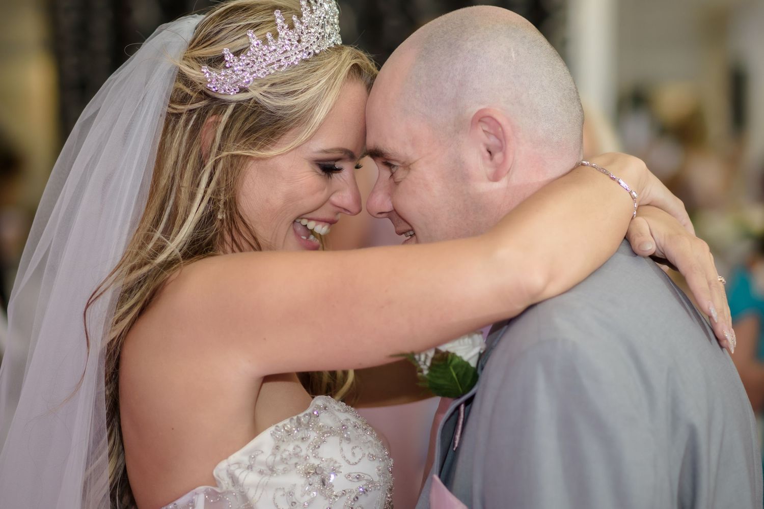 Bride in tiara and veil embraces bald groom, both smiling joyfully at their wedding reception.