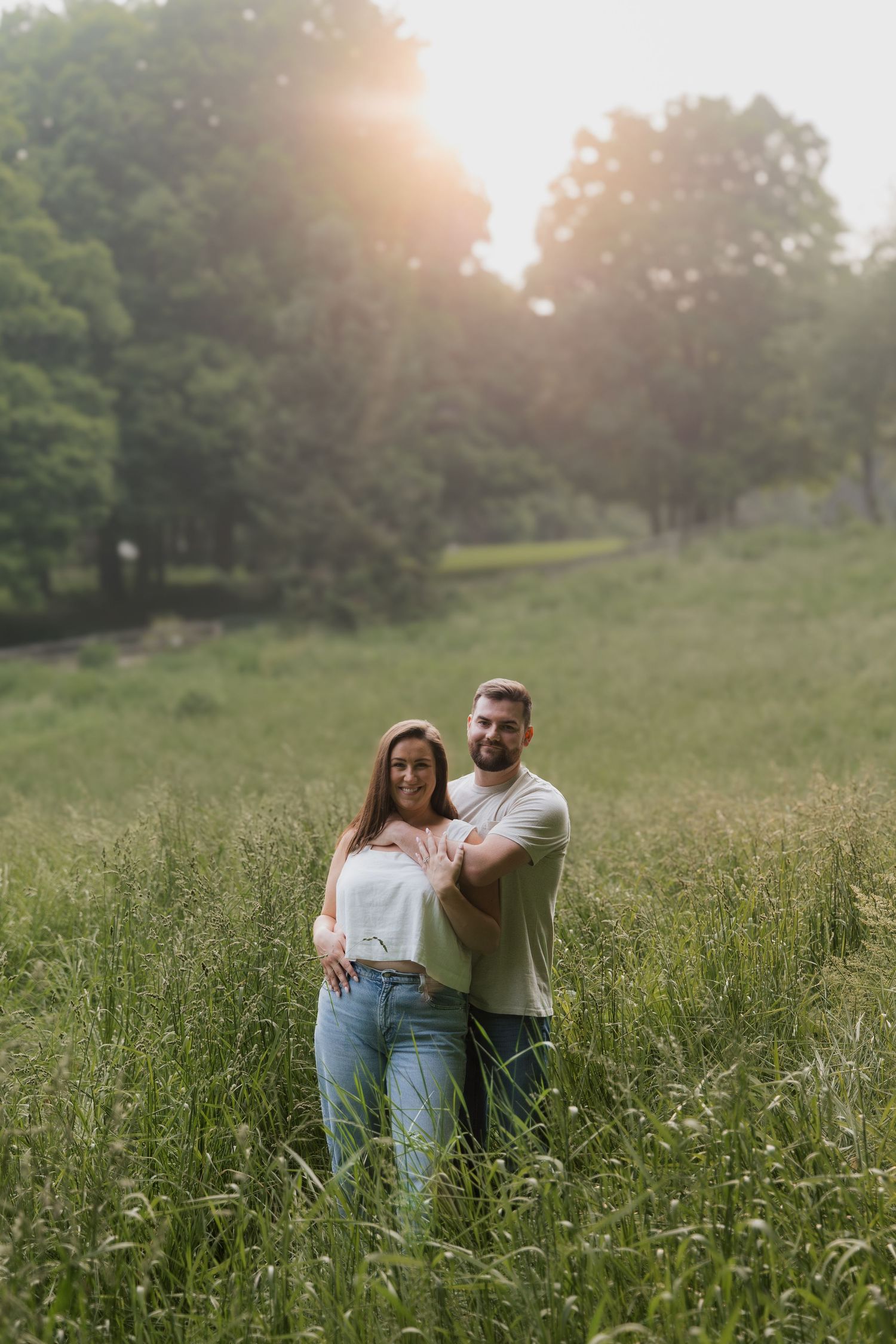 A smiling couple embraces in a sunlit green meadow during a golden hour engagement photo session.
