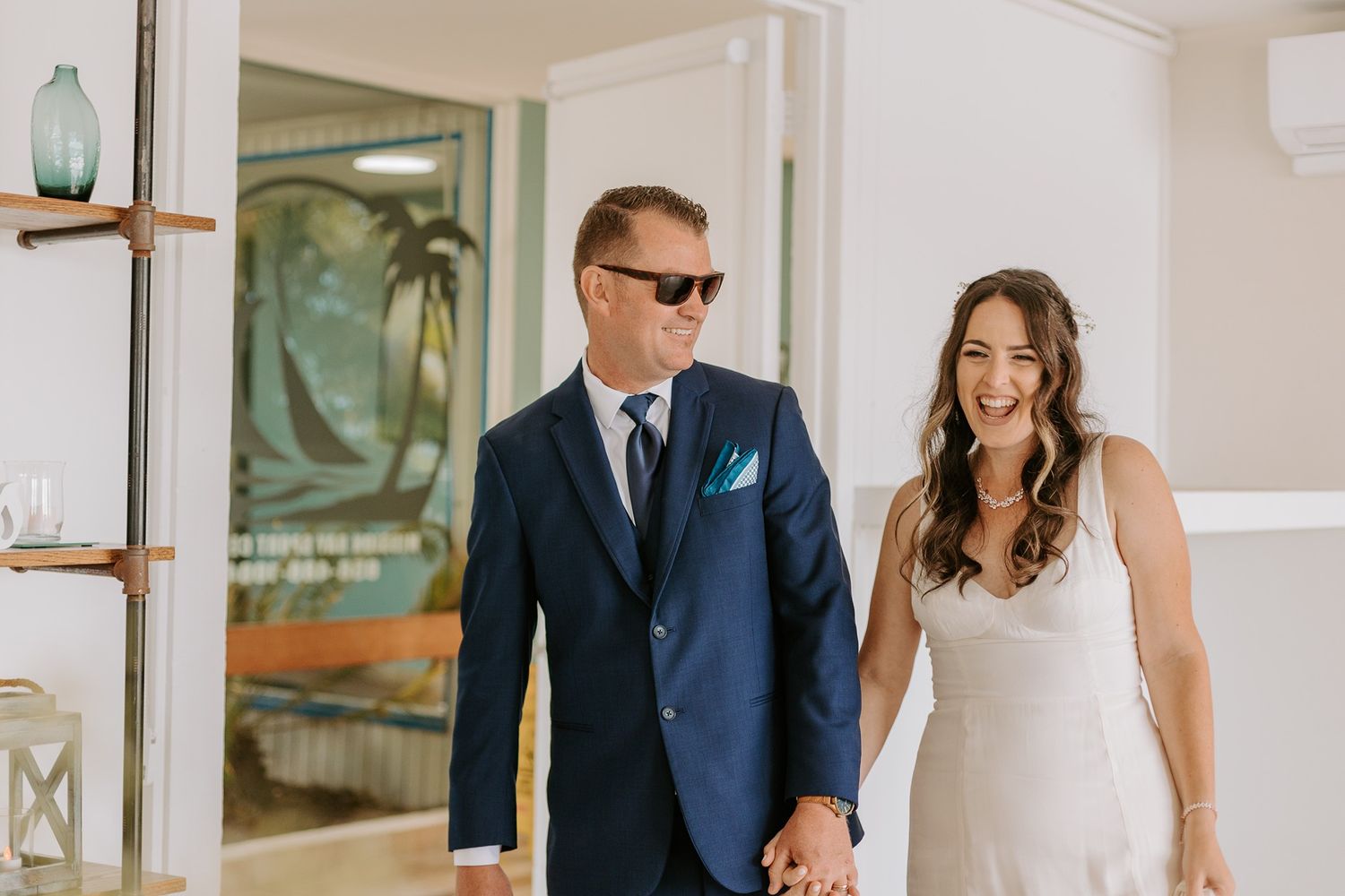 Happy couple in wedding attire holding hands, groom in navy suit with sunglasses, bride in white dress, smiling indoors at Tower Beach Club.