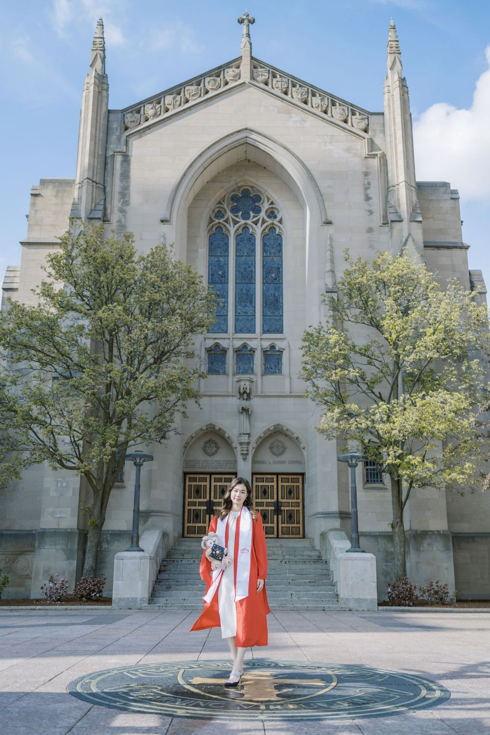 Boston University Graduate in red gown stands before grand Gothic university chapel with ornate stained glass windows on sunny day.