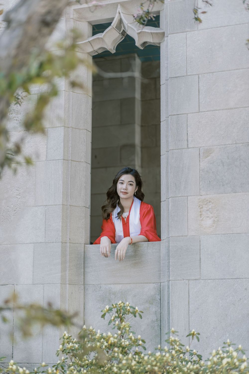 Boston University graduate in a red robes leans out of a stone building window, surrounded by lush greenery.