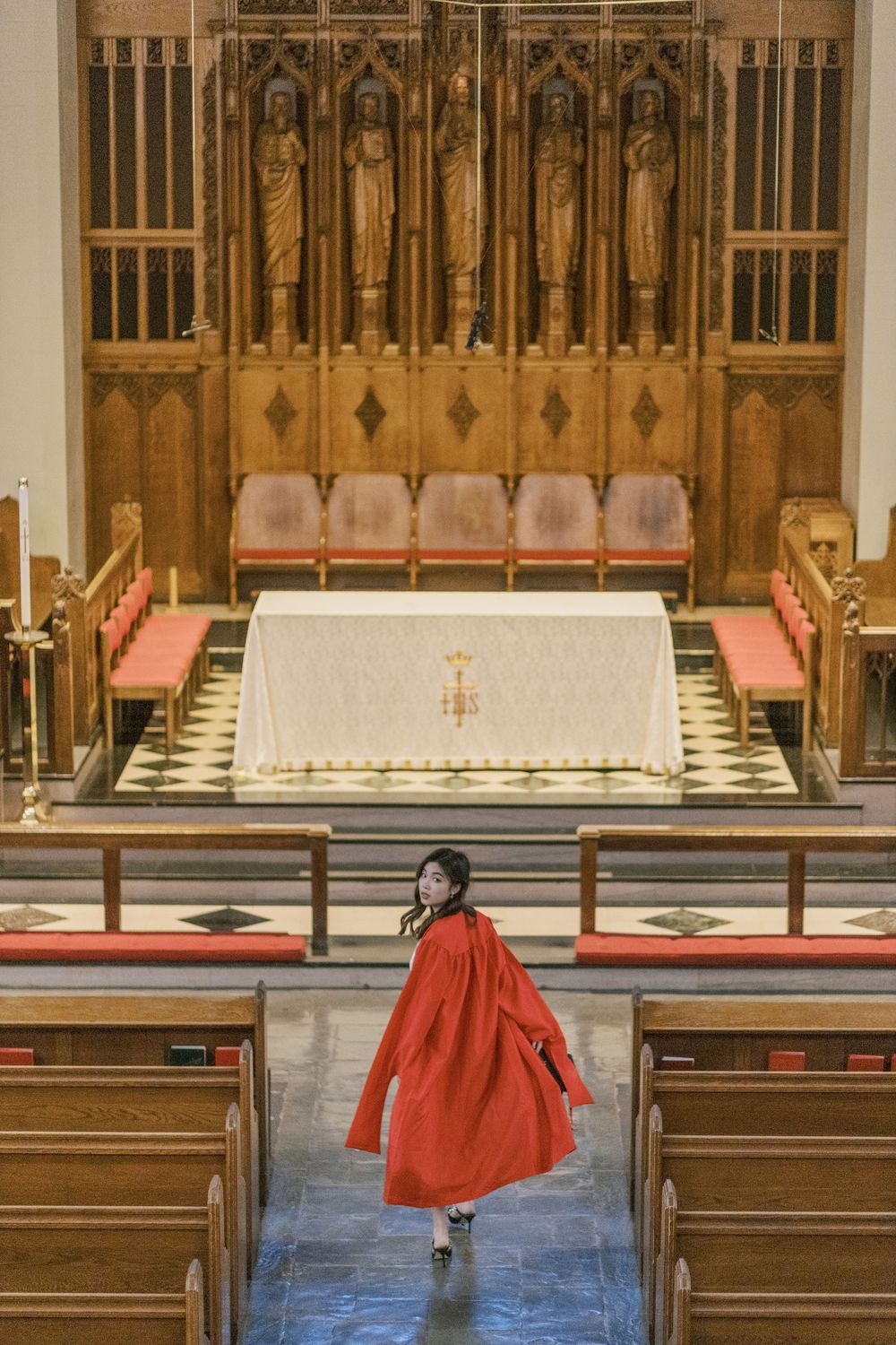 Boston University graduate in red coat standing in church aisle before ornate wooden altar with religious statues.