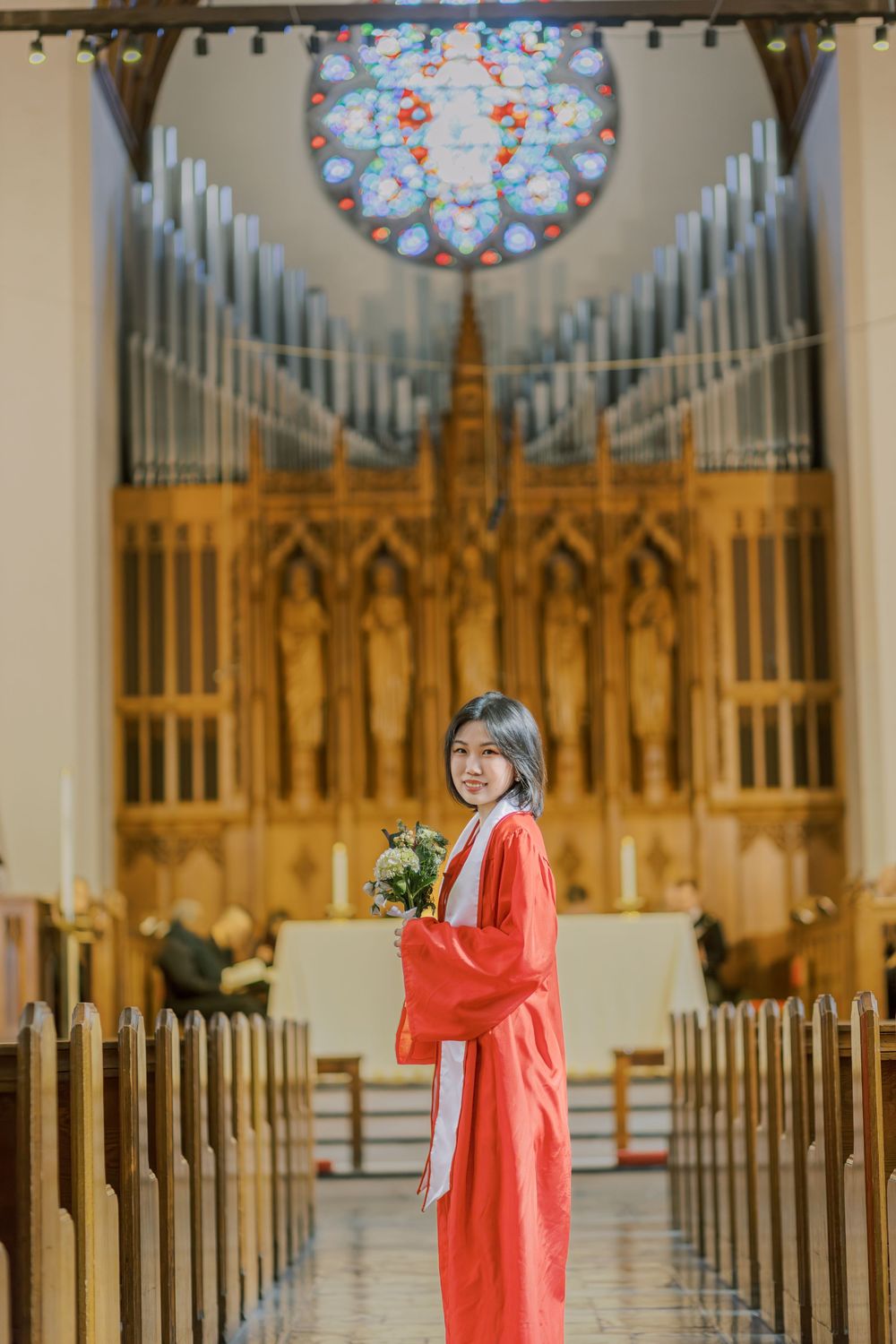 Young woman in red Boston University graduation gown holding flowers smiling inside a church with stained glass window.
