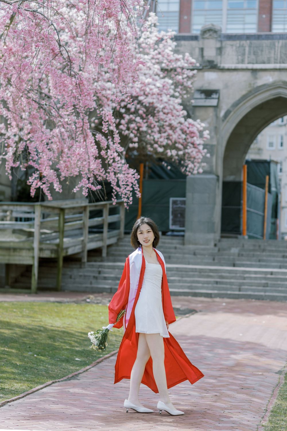 Boston University graduate in white dress and red kimono walks under pink cherry blossoms near stone building.