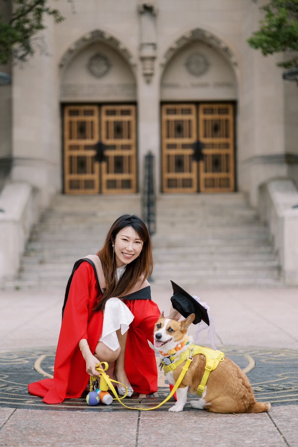 Boston University Graduate in red gown poses with Corgi wearing graduation cap outside university building.