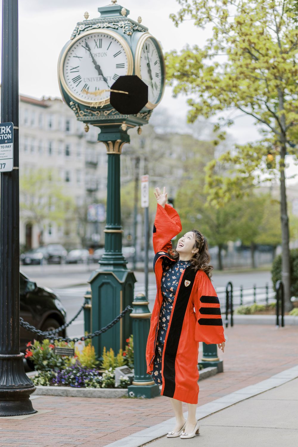 Graduate in red Boston University doctoral robe celebrates near a classic street clock on a brick sidewalk in an urban setting.
