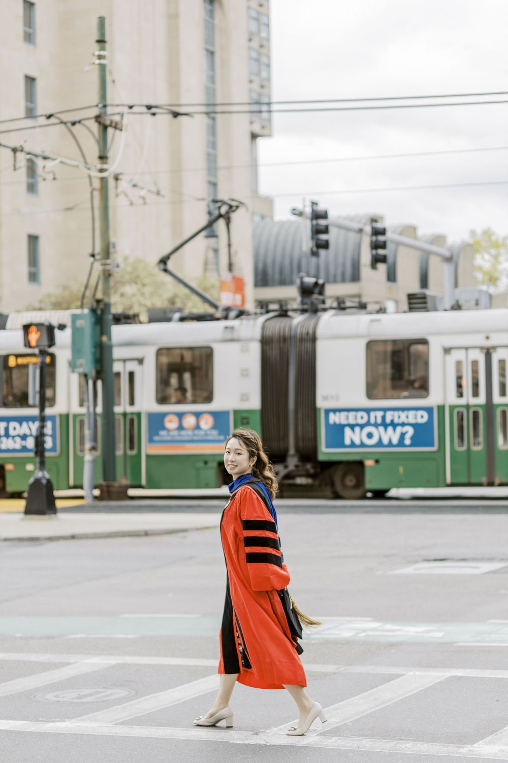 Woman in red doctoral Boston University graduation gown crossing street with green MBTA trolley in Boston, MA.
