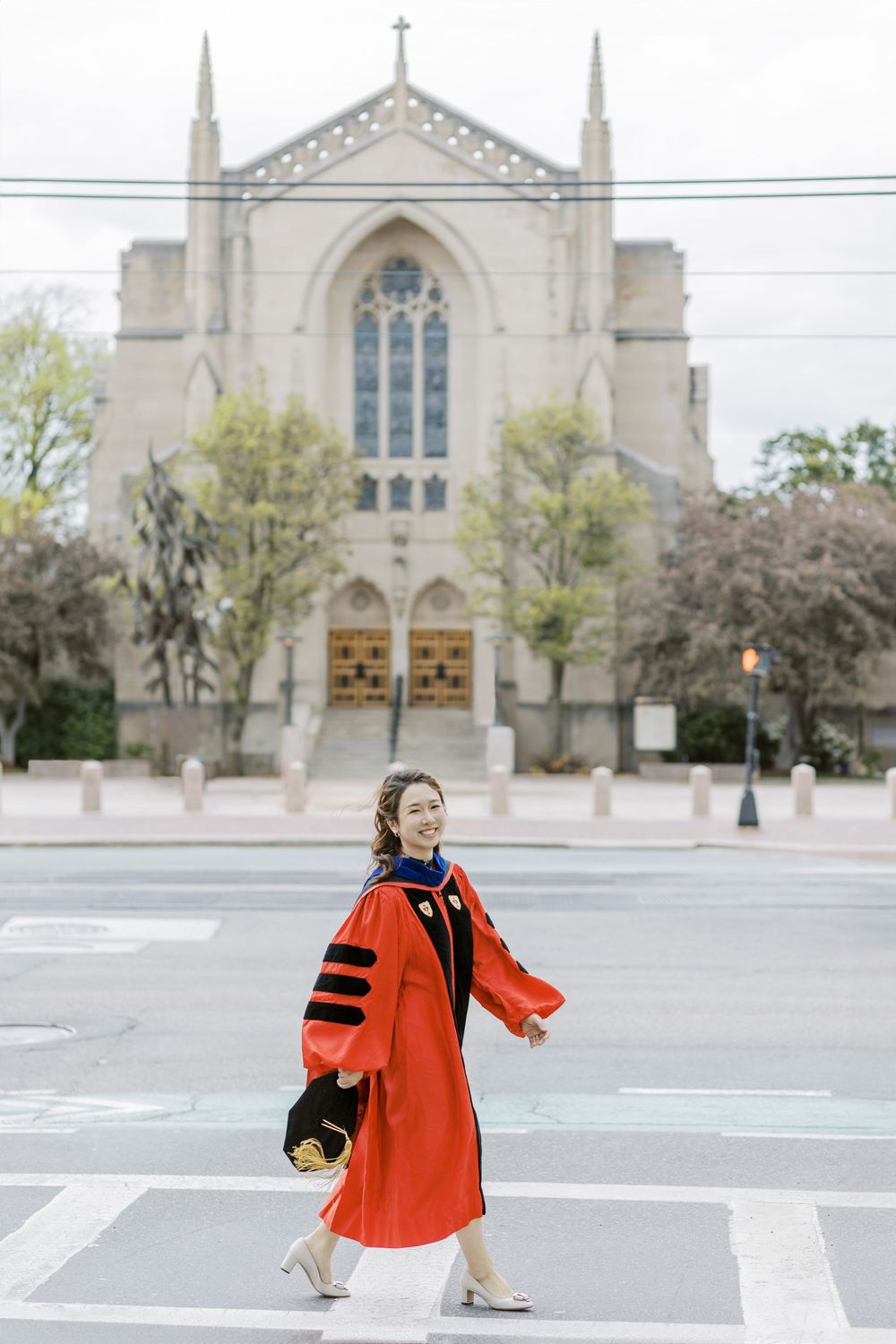 Woman in red doctoral Boston University graduation gown crossing street in front of gothic university chapel.