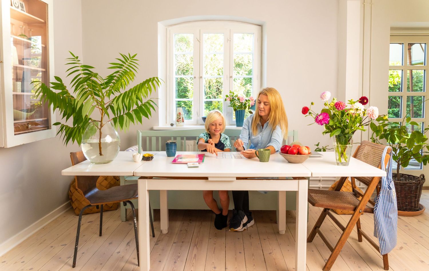 Two women sit at a white farmhouse dining table surrounded by lush plants and fresh flowers in a bright Scandinavian home.