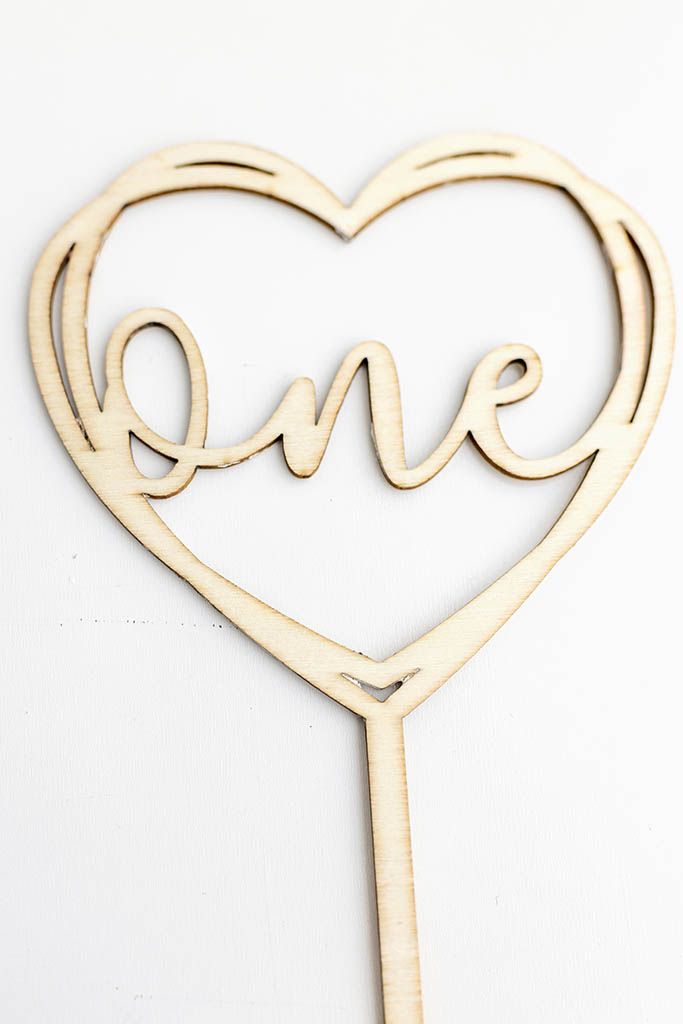 Wooden laser cut heart shaped cake topper with cursive word one on white background.