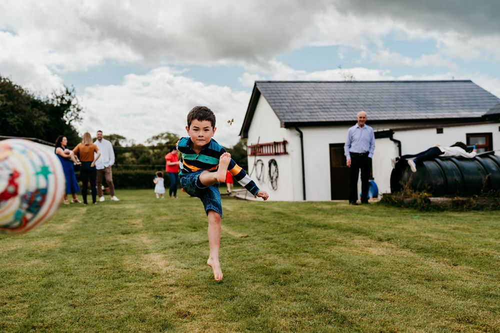 A barefoot boy kicks a soccer ball on a green lawn with a white cottage and people in the background.
