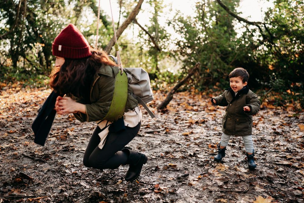 Woman with backpack and red hat playing with toddler boy in autumn forest leaves outdoor family fun.