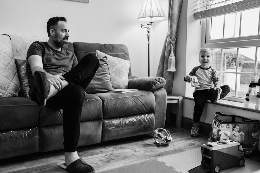 Black and white photo of a man relaxing on a sofa while a young child plays nearby in a cozy living room.