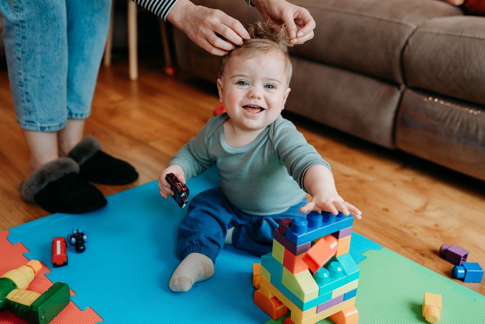Happy baby smiling while playing with colorful building blocks on a blue mat as an adult styles their hair.
