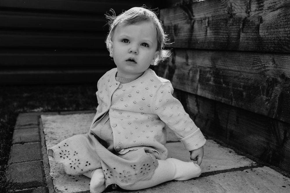 Black and white photo of a cute baby girl sitting outdoors on a patio near a wooden fence.