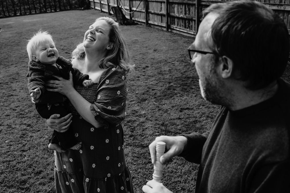 Black and white photo of a laughing woman holding a baby while a man smiles nearby in a garden.