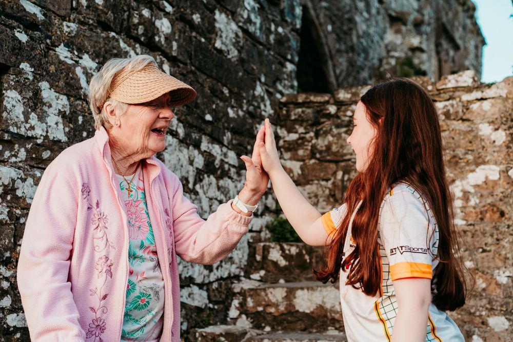 Elderly woman and young girl high-fiving outdoors near a stone wall, sharing a joyful intergenerational moment.