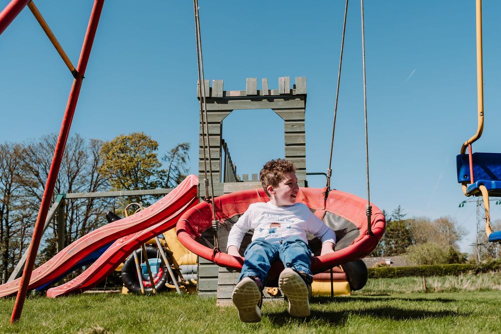 Child swinging on a red nest swing in a sunny playground with a slide and castle-shaped frame.