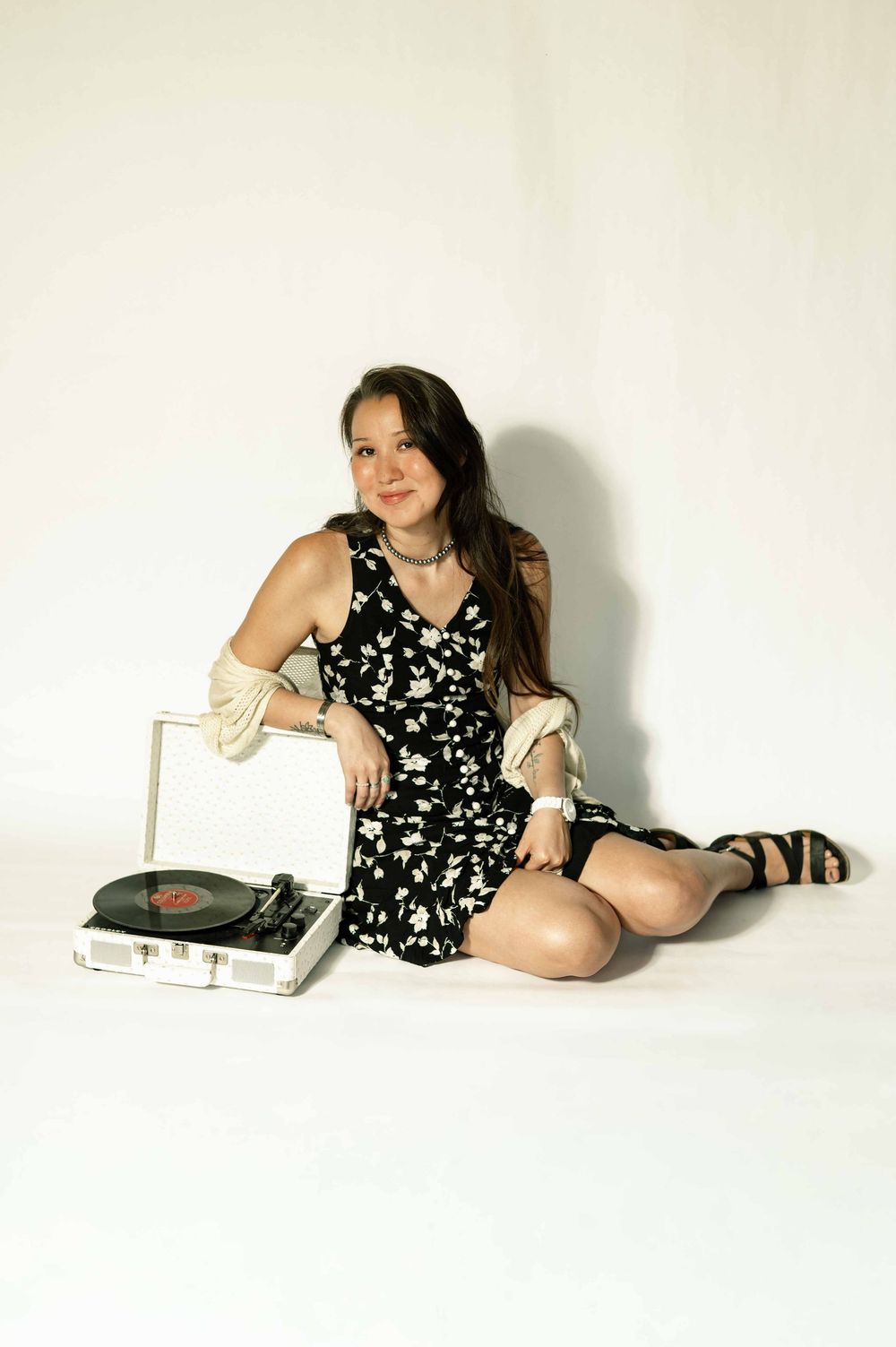Young woman in floral dress sitting on floor with vintage record player, smiling, long brown hair, white background.