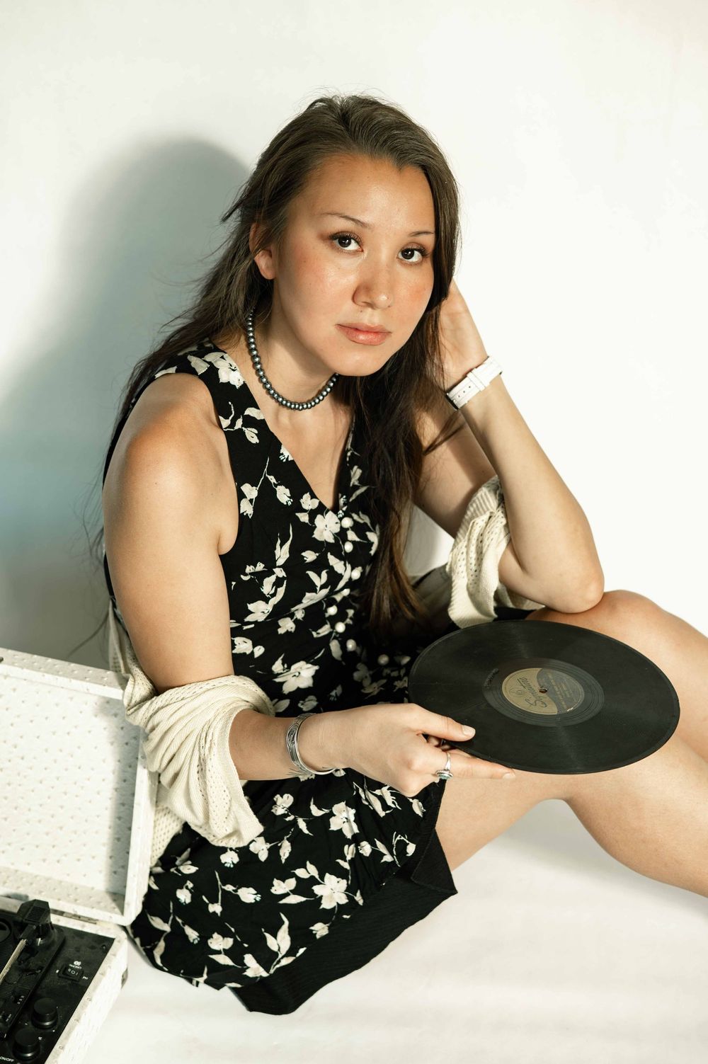 Young woman in black floral dress sitting on floor holding vinyl record with a case beside her.