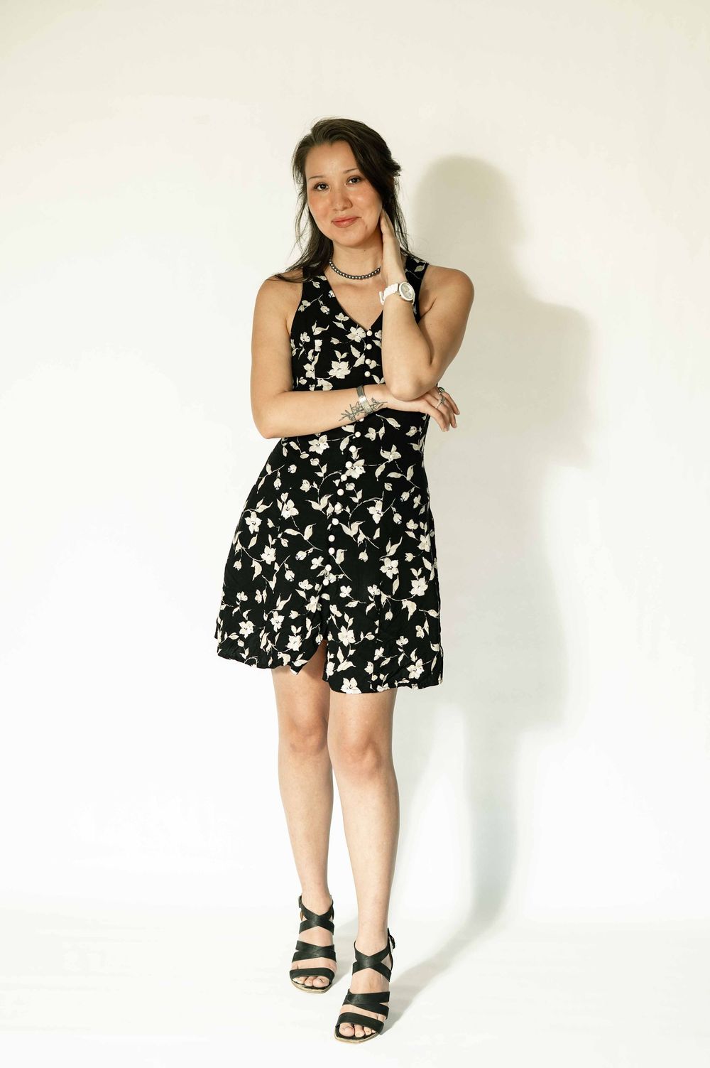 Young woman in black floral mini dress and strappy heels posing against white background.
