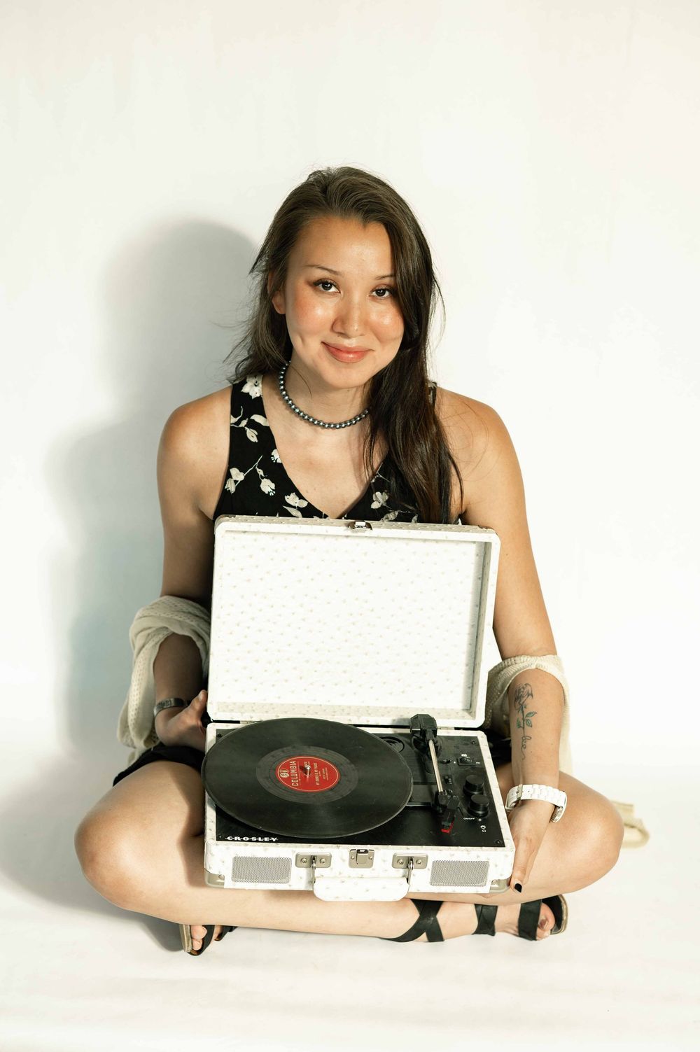 Young woman sitting cross-legged holding a white vintage record player with a vinyl record on white background.