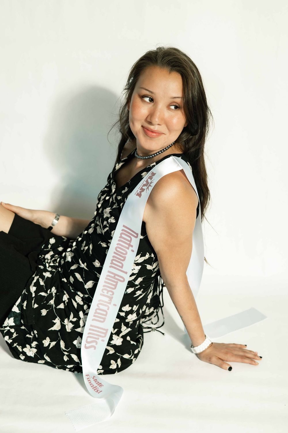 Young woman wearing a National American Miss pageant sash, sitting gracefully in a floral dress against a white background.