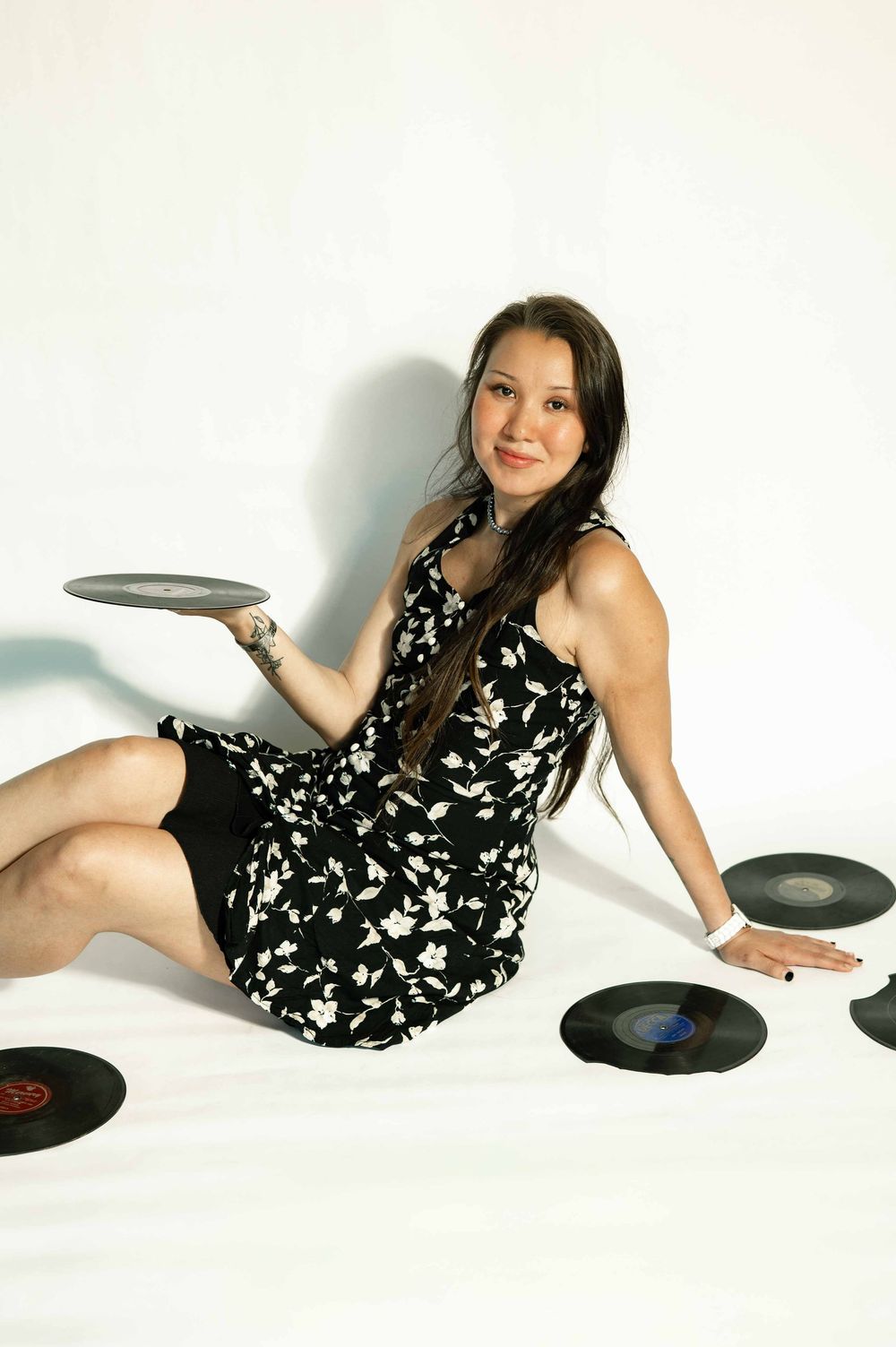 Smiling young woman in black floral dress sitting on floor surrounded by vinyl records.