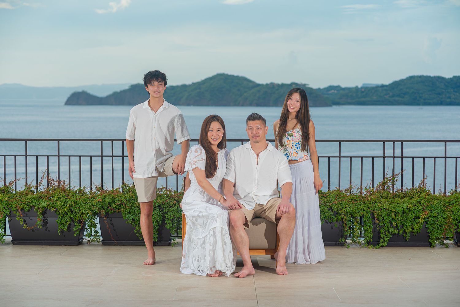 Family of four posing on a scenic ocean-view terrace at sunset, two adults and two boys in casual summer attire.