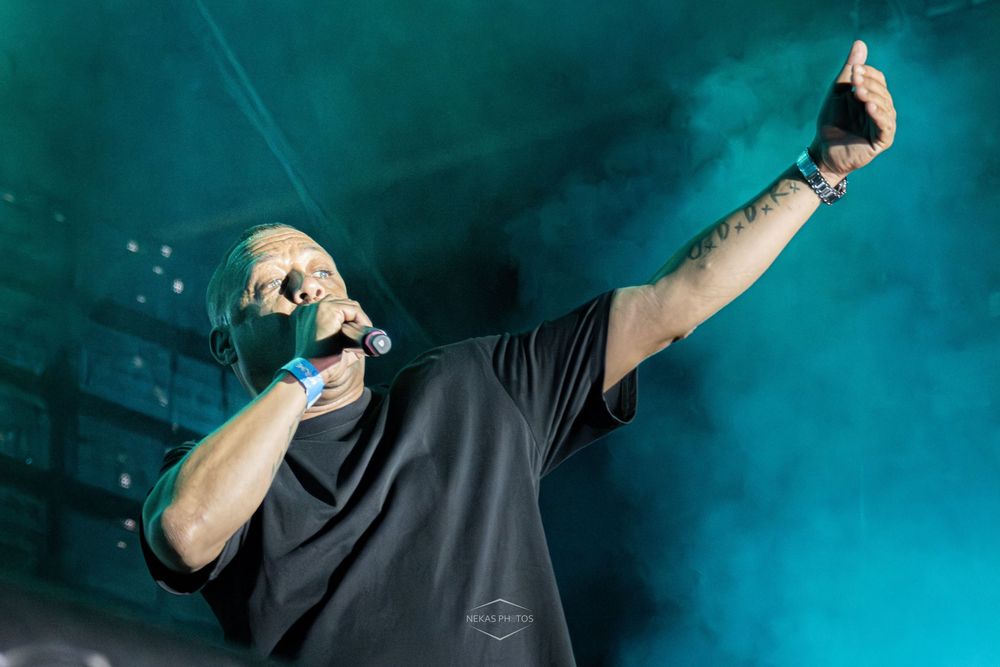 Rapper performing on stage with microphone, raising hand under teal smoke lights at a live concert.