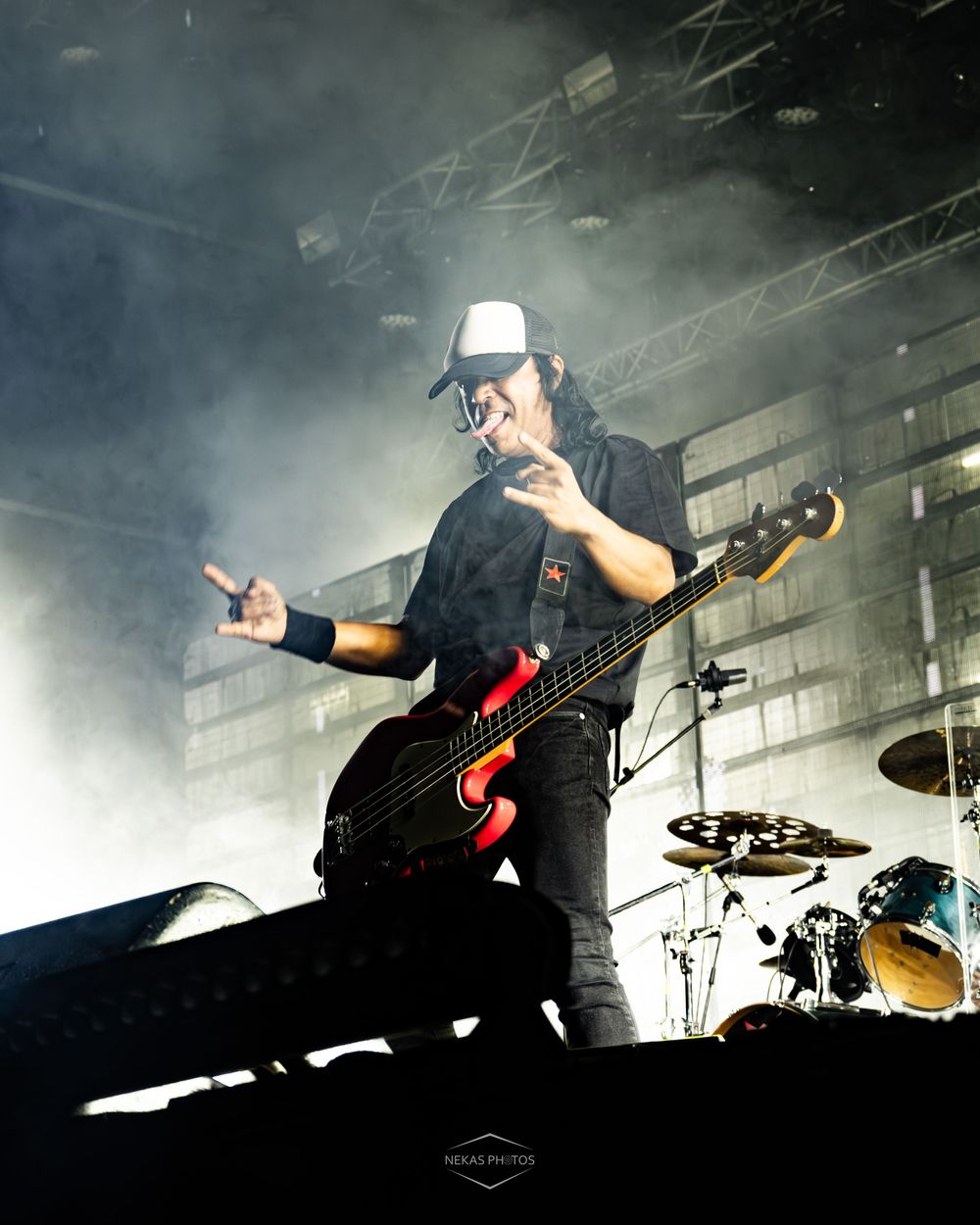 A guitarist performs on stage with a bass guitar, wearing a white cap, amid dramatic fog and stage lighting.
