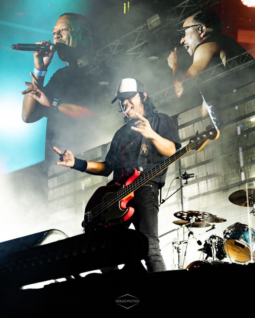 Rock band performing live on stage with bassist in foreground and two vocalists in background amid dramatic fog lighting.