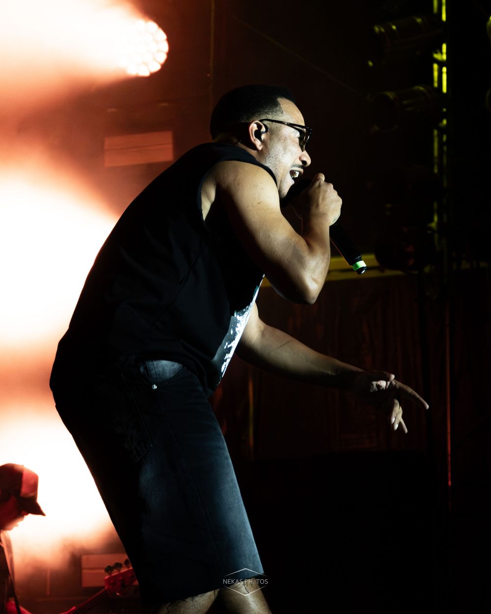Male rock vocalist performing on stage, leaning forward with microphone, wearing black sleeveless shirt under dramatic concert lighting.