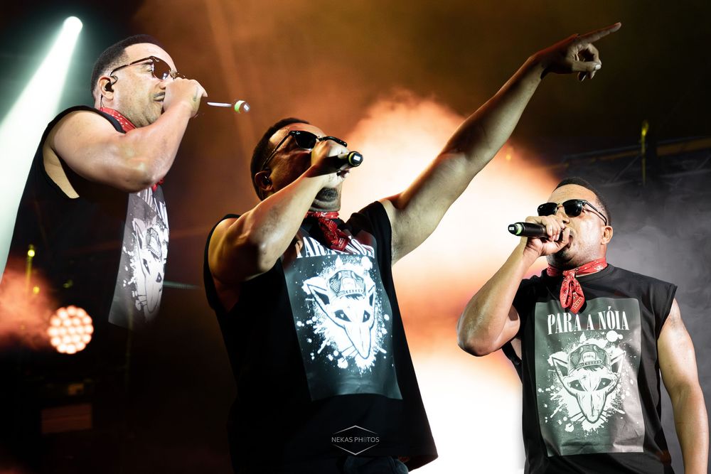 Three male hip-hop artists performing on stage with microphones under dramatic orange stage lighting.