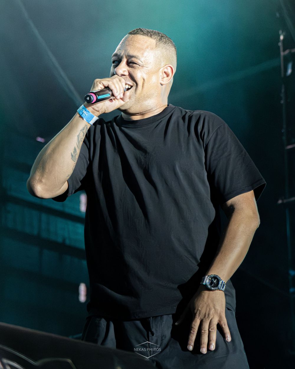 A man in a black t-shirt performs on stage, smiling and holding a microphone under dramatic blue stage lighting.
